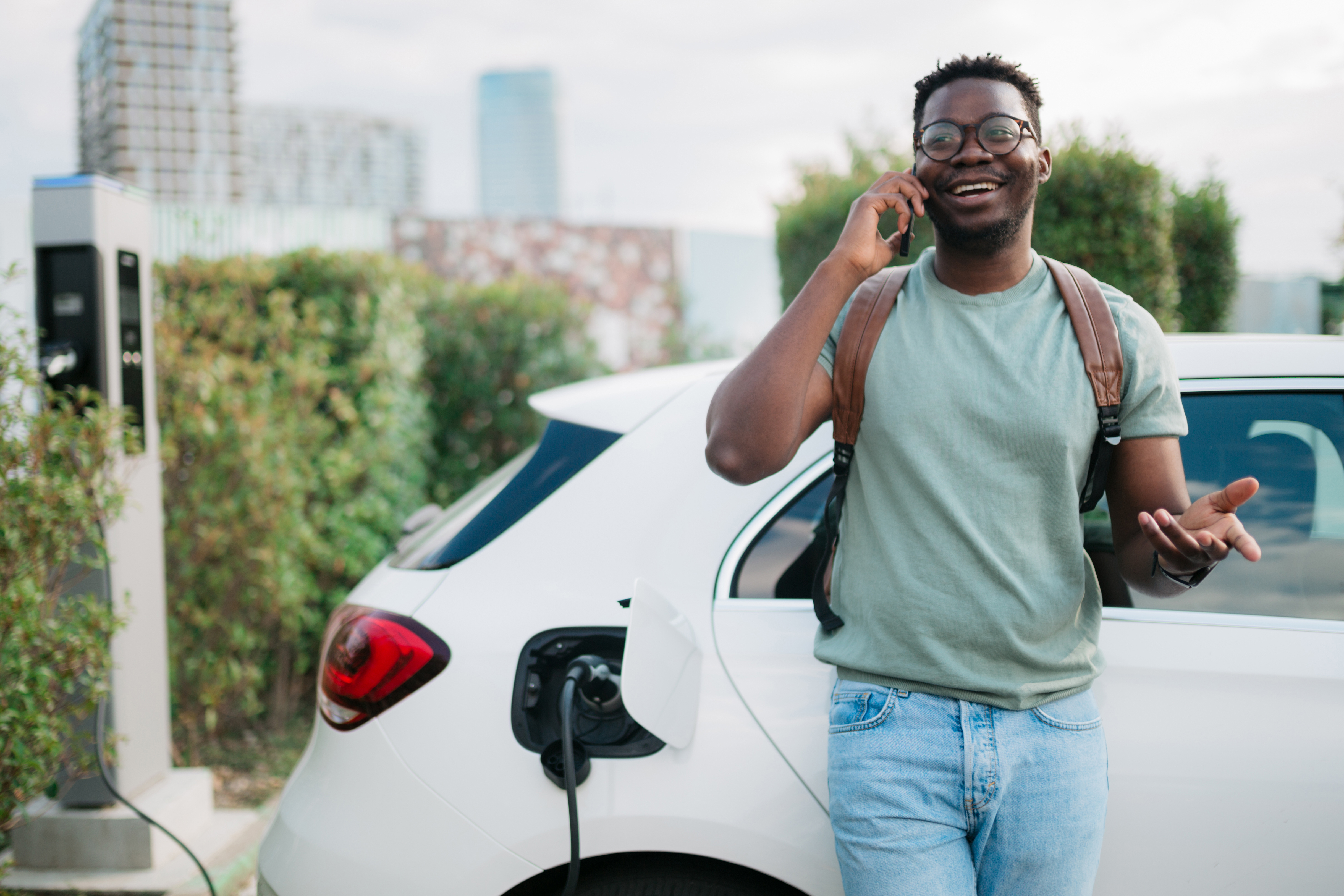 Happy young adult standing next to his electric vehicle and using his smart phone while his car is plugged into the charging station